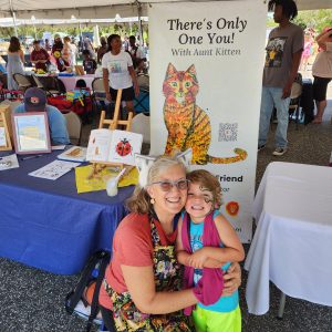 Kristin hugging great-nephew in front of her book table at a free book giveaway event. There is a large banner behind them saying "There's Only One You! With Aunt Kitten"