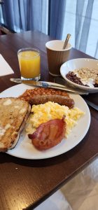 Breakfast of scrambled eggs, sausage, bacon, baked beans, toast, fruit with yogurt and granola, Orange juice, and latte